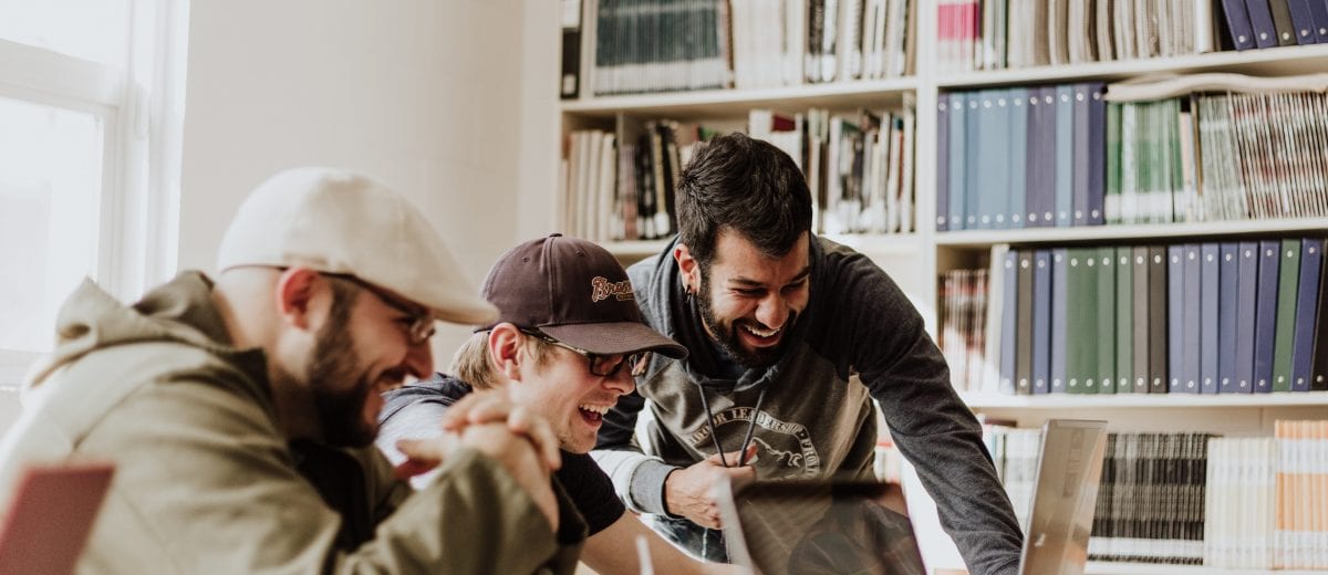 photo of three men looking at laptop and laughing in library