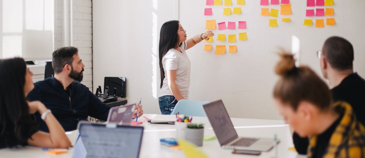 photo of woman talking to a group of people at a table in front of a wall covered in sticky notes